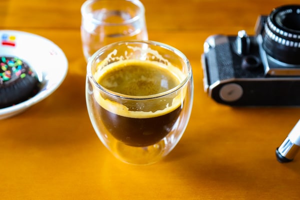 How Much Caffeine in Decaf Espresso: Brewing Insights 8 A clear double-walled glass filled with decaf espresso, placed on a wooden table beside a dessert and camera