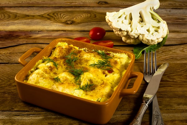 Cauliflower Bake: Deliciously Cheesy and Healthy Recipe 8 A golden-baked cauliflower casserole, topped with fresh dill, served in an orange dish with a side of cherry tomatoes and a cauliflower head on a wooden table