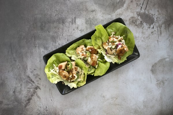 21 Keto Chinese Recipes: Enjoy Takeout Without the Carbs 9 A rectangular black plate holds three lettuce wraps filled with meat and vegetables, placed on a gray stone surface.