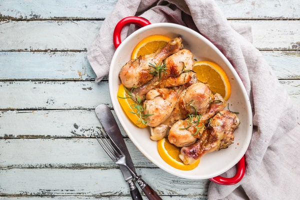 13 Keto Crockpot Chicken Recipes for Effortless Cooking 2 A top-down view of six baked chicken drumsticks garnished with fresh rosemary and orange slices in a white and red oval dish on a rustic light-blue wooden surface.