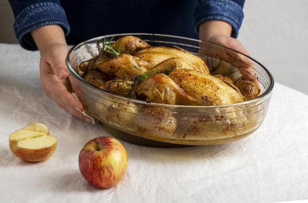 13 Keto Crockpot Chicken Recipes for Effortless Cooking 5 A person in a denim shirt holds a large, oval glass baking dish filled with two roasted chickens and potatoes, set on a white tablecloth next to a whole and a halved apple.