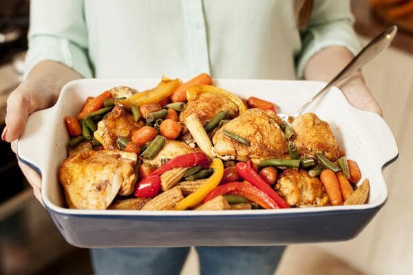 13 Keto Crockpot Chicken Recipes for Effortless Cooking 9 A person wearing a light green shirt holds a white and dark blue baking dish filled with roasted chicken pieces, baby corn, green beans, carrots, and colorful bell peppers.