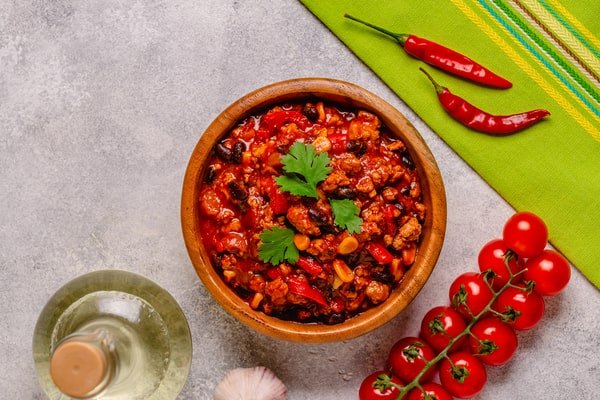 Keto Turkey Chili With Big Flavor Boost 3 A wooden bowl of keto turkey chili garnished with fresh parsley, surrounded by cherry tomatoes, chili peppers, and a bottle of oil
