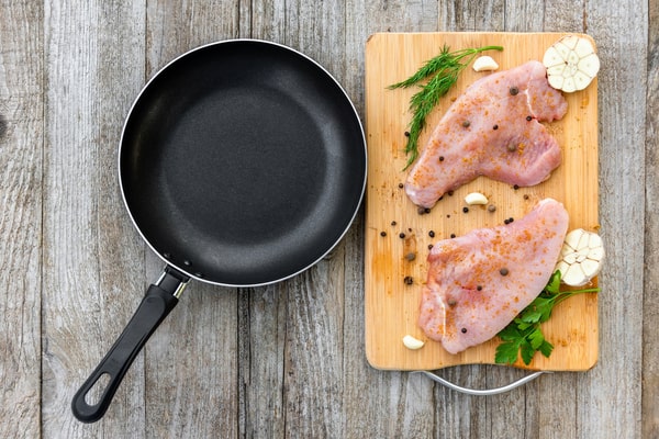 Pan Seared Chicken Breast Made Easy in Just Minutes 4 Raw seasoned chicken breasts on a wooden board with herbs, garlic, and a frying pan beside them