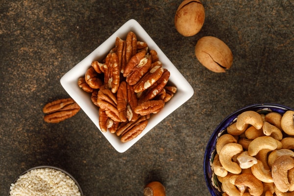 A square white bowl filled with pecans beside cashews and other nuts on a dark tabletop
