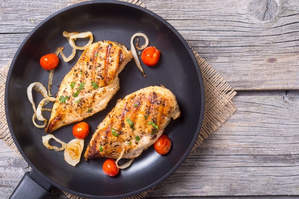 Pan Seared Chicken Breast Made Easy in Just Minutes 7 Two chicken breasts in a pan with onions and cherry tomatoes on a wooden surface
