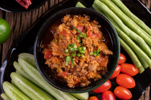 Keto Turkey Chili With Big Flavor Boost 7 A bowl of keto turkey chili garnished with chopped green onions, surrounded by fresh cucumber sticks, long beans, and cherry tomatoes on a black plate
