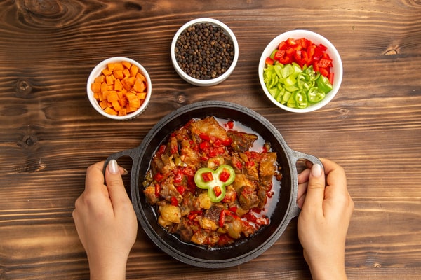Keto Turkey Chili With Big Flavor Boost 10 A skillet of keto turkey chili with chopped peppers and carrots, held by two hands, surrounded by bowls of colorful vegetables and spices on a wooden table