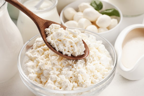Low Carb Cottage Cheese: Best Brands and Ways to Enjoy 3 A wooden spoon is held over a clear glass bowl overflowing with lumpy cottage cheese, surrounded by various other dairy products like milk, yogurt, and small balls of mozzarella.