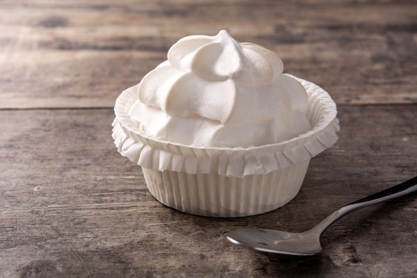 Is Cool Whip Low Carb? A Guide for Low-Carb Dieters 4 A white cupcake wrapper holds a tall, swirled meringue or whipped cream topping next to a small silver spoon on a rustic wooden table.