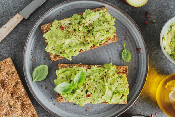 An overhead view shows two slices of crispbread topped with mashed avocado, seasoning, and basil leaves on a dark grey plate next to a knife and bottle of oil.
